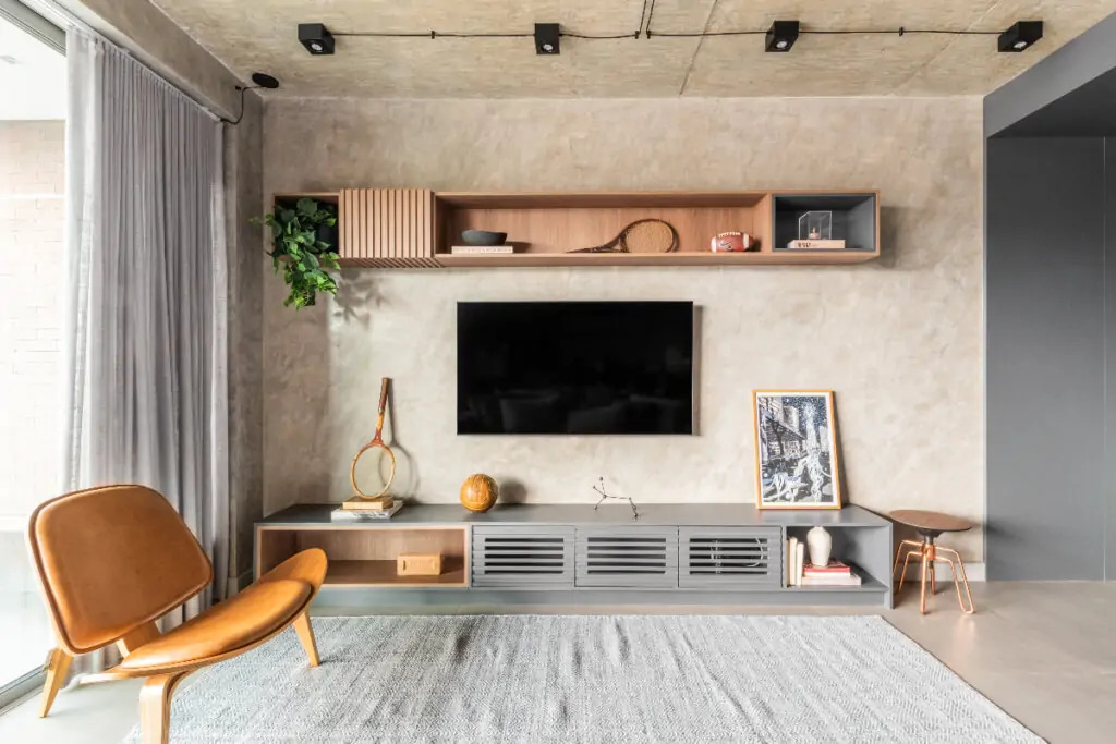 Imagem de sala com cadeira marrom, tapete cinza, tv na parede e prateleira acima dela com objetos 