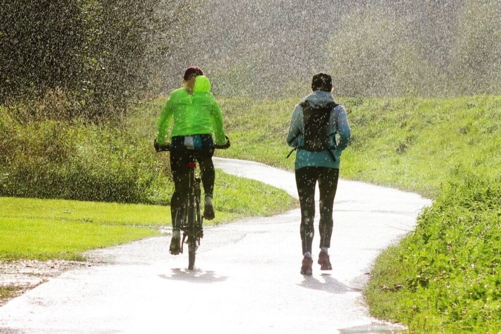 Mulher com blusa verde neon pedalando ao lado de outra mulher correndo, ambas na chuva 