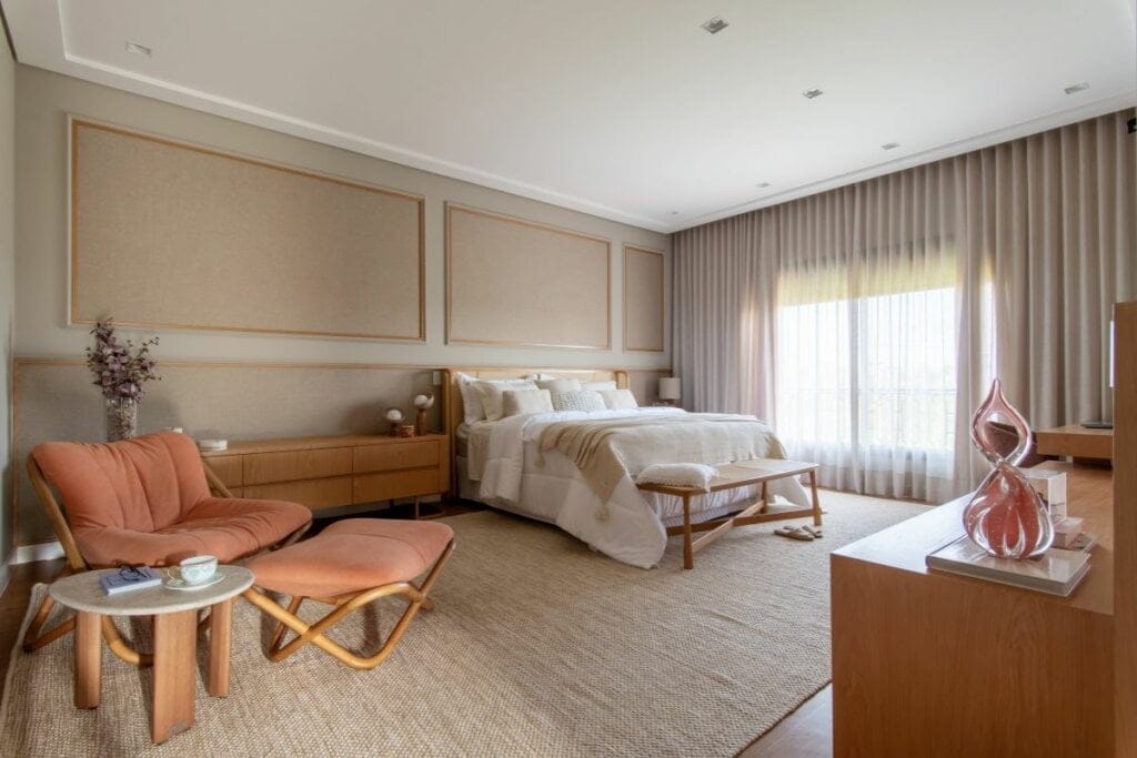 Dormitório com cama com forro branco e bege, tapete bege e janela lateral com cortinas bege 