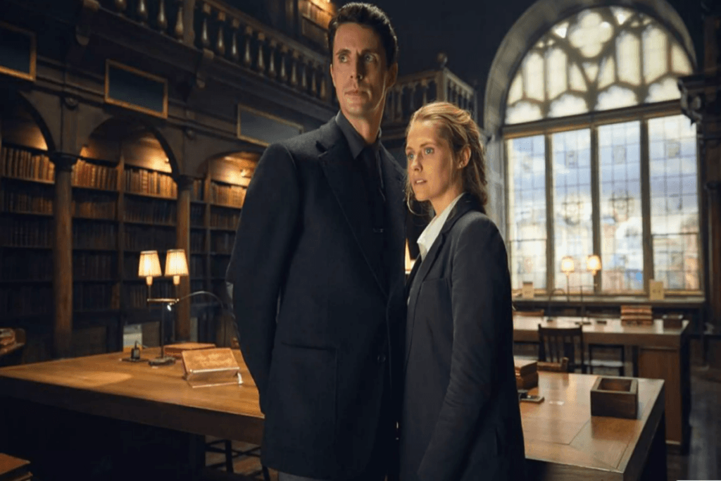 Imagem de um homem e uma mulher encostados um no outro em biblioteca, os dois vestem roupas formais