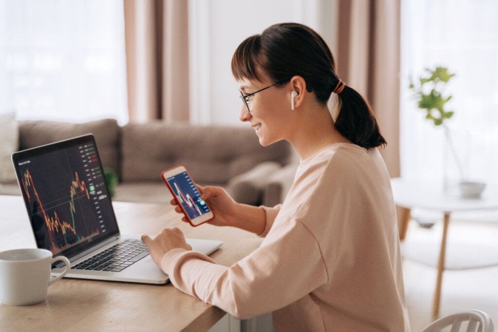 Mulher sentada em casa, usando óculos e fones de ouvido, analisando gráficos no notebook enquanto segura o celular com aplicativo de investimentos.