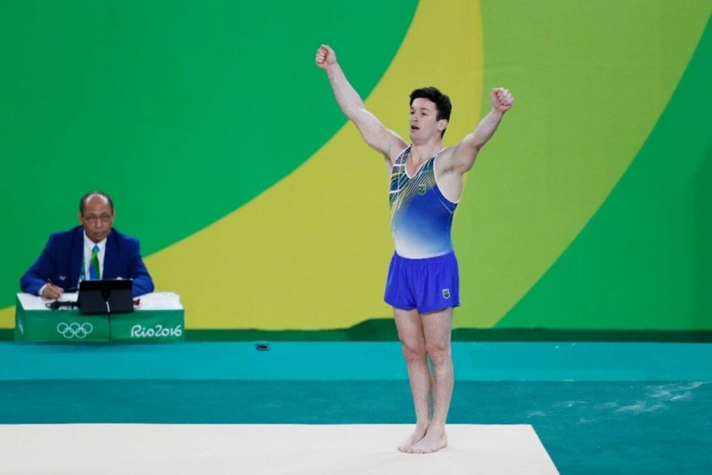 Homem com roupa de ginástica com os braços para cima em tablado de competição