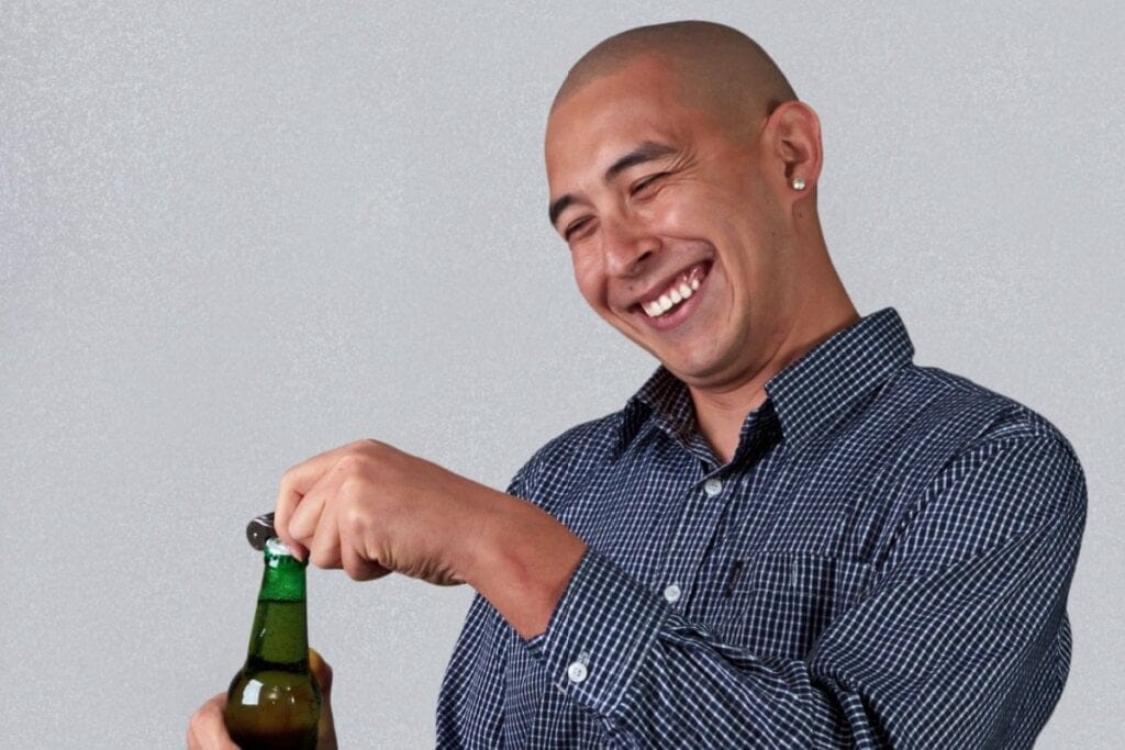 homem careca de camisa azul sorrindo e abrindo garrafa de vidro com abridor