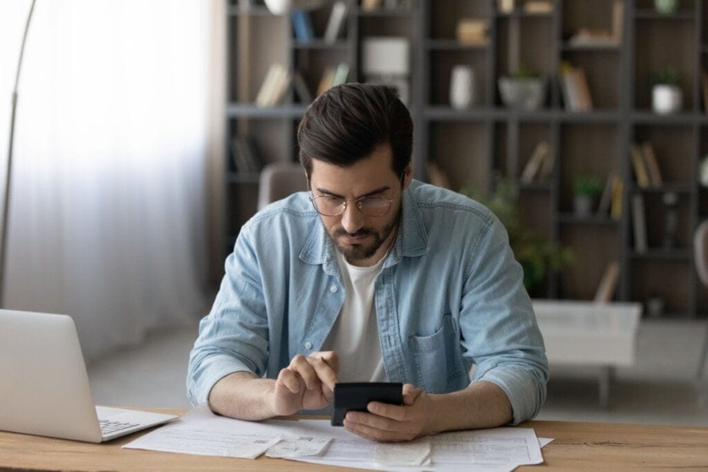 Homem usando camisa azul e camiseta branca, óculos e com o cabelo curto com uma calculadora na mão sentado em mesa com contas