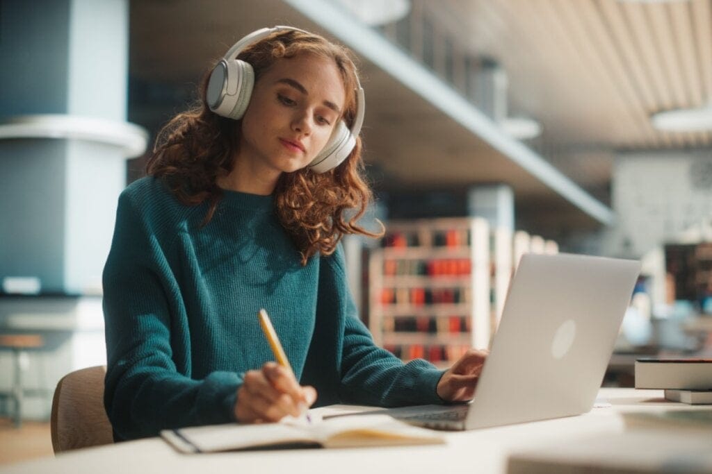 Jovem usando blusa de frio verde e com fones de ouvido branco escrevendo em caderno com notebook na frente