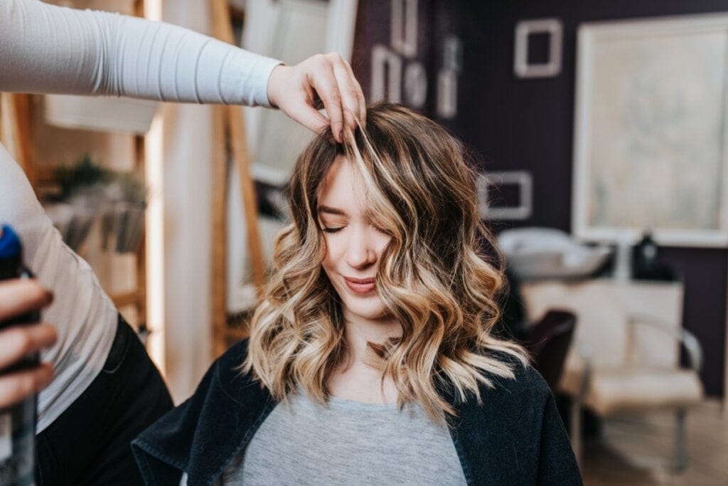 Mulher com cabelo com mechas loiras sentada e uma mão mexendo em seu cabelo 