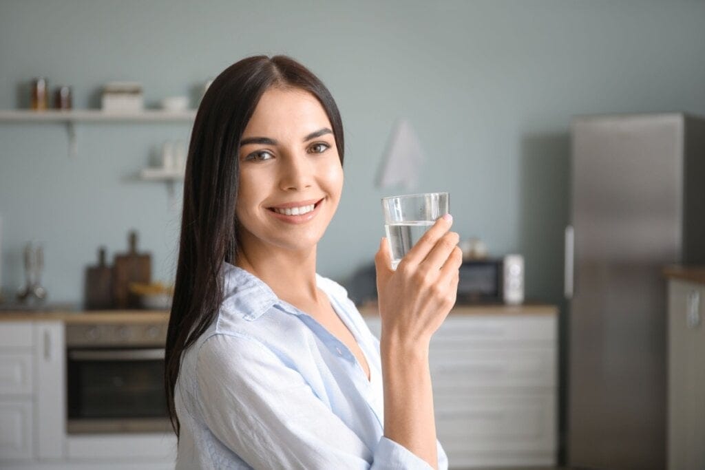 Mulher com cabelo liso preto e solto, usando uma camisa azul clara, com um copo de água na mão e sorrindo