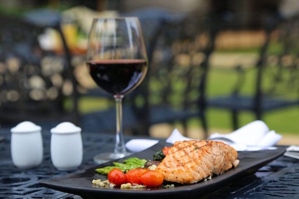 Salmão grelhado servido em prato quadrado preto, junto com tomate-cereja e aspargos. Ao lado uma taça de vinho Pinot Noir em cima de mesa de madeira