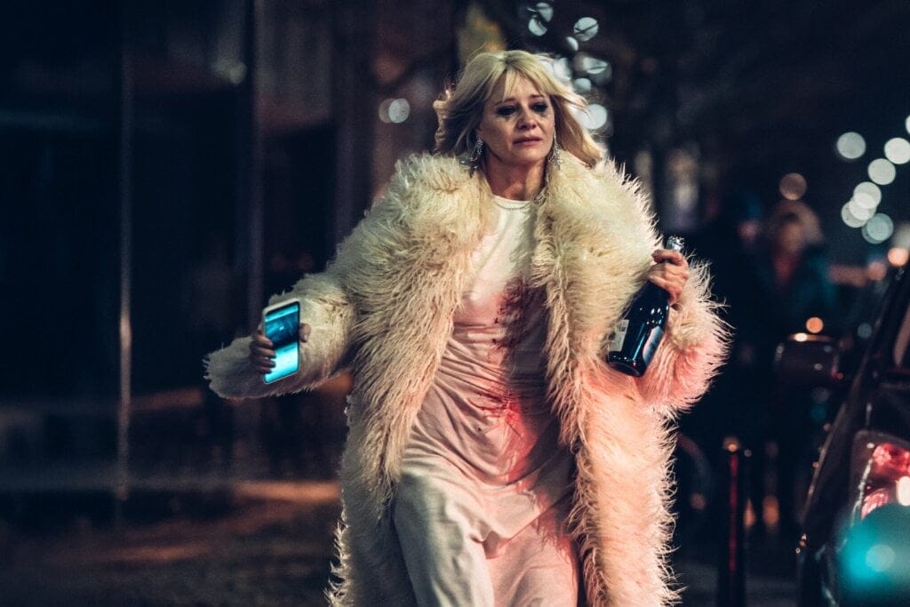 Mulher usando um casaco peludo e vestido, andando na rua, segurando o celular e uma garrafa de champanhe