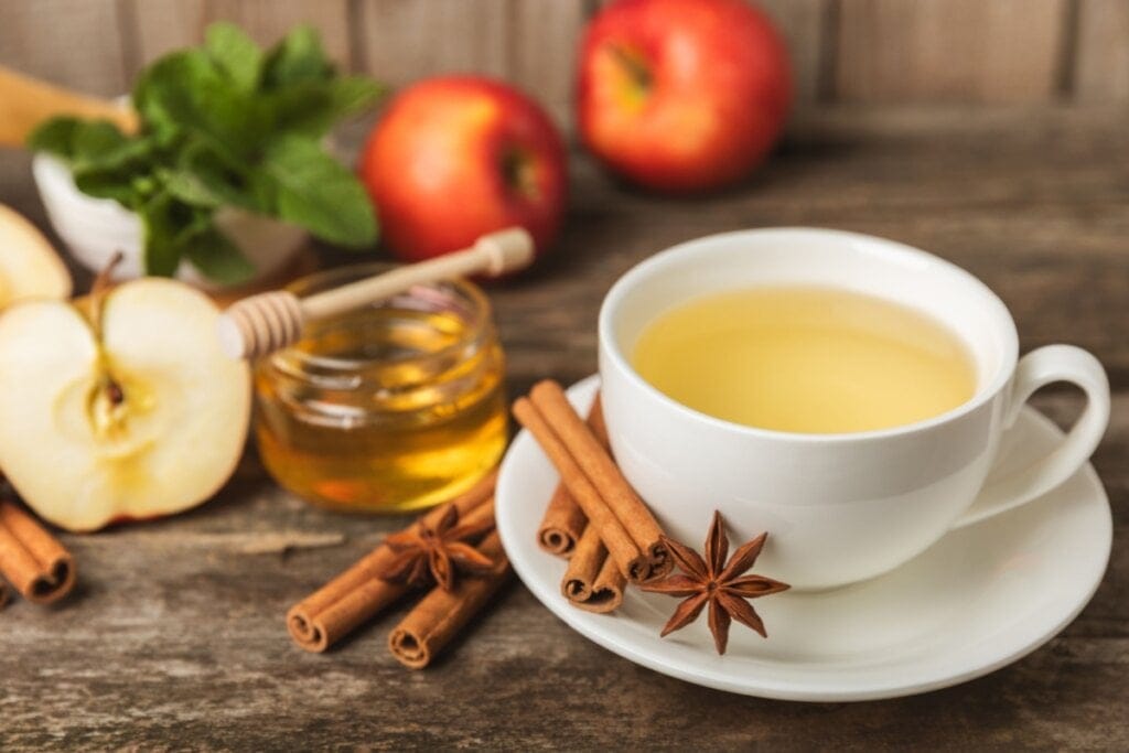 xícara branca com chá amarelo em cima de pires branco com canela e anis-estrelado. maçãs e pote com mel ao fundo