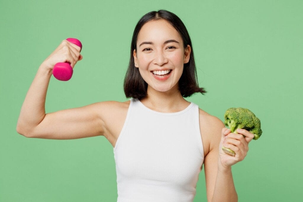 Jovem com cabelo preto curto, usando uma regata branca e em uma mão há um peso de academia rosa e na outra um brócolis em fundo verde