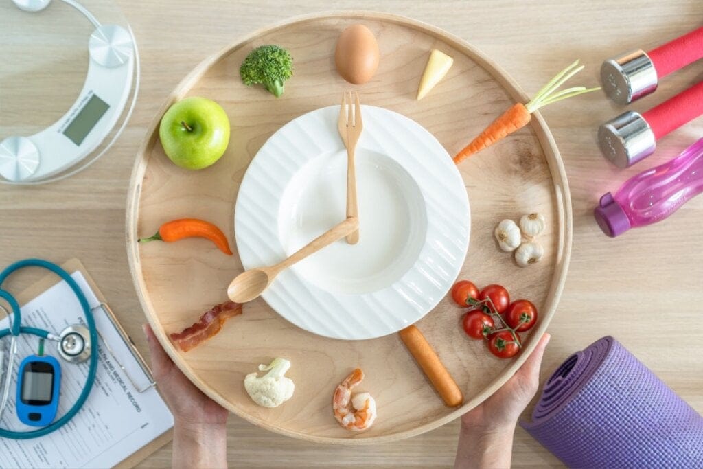 prato com talheres imitando um relógio e alimentos saudáveis em volta, como ovo, maçã, tomate etc.