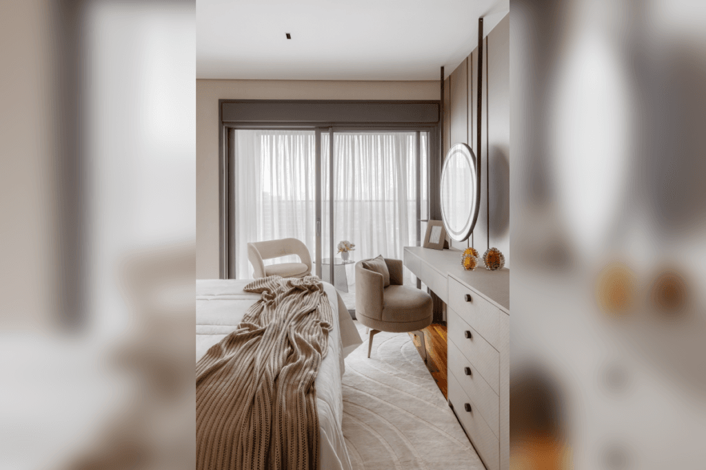 Quarto com cama coberta por manta de tricô bege, tapete claro e poltrona próxima à escrivaninha com espelho redondo iluminado