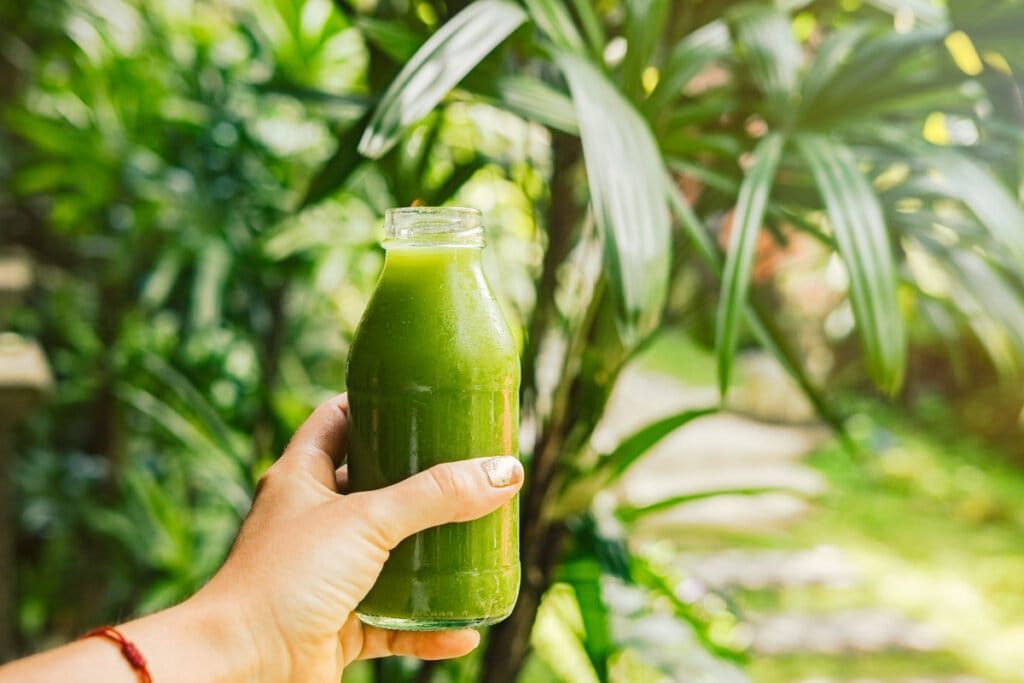 Mão feminina segurando uma garrafa com suco verde em frente a uma árvore