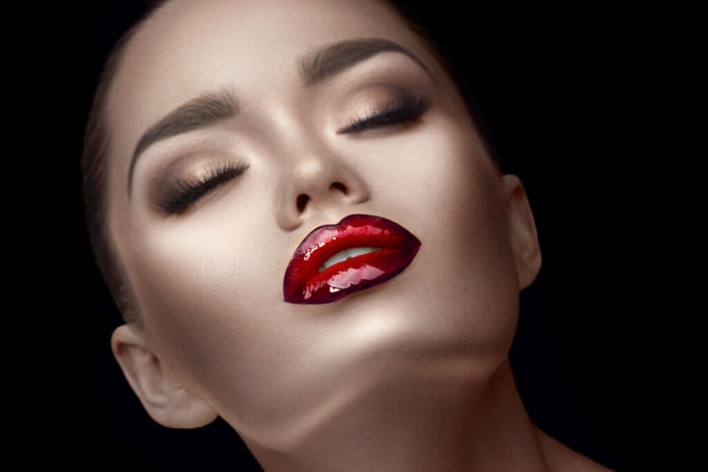 Close-up de um rosto feminino com maquiagem intensa, olhos fechados, batom vermelho brilhante e expressão serena