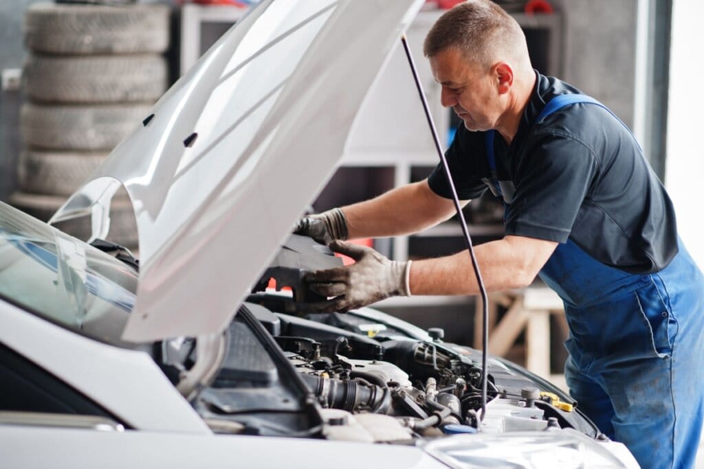Homem com camiseta azul-escura, usando macacão jeans fazendo manutenção em carro branco na oficina