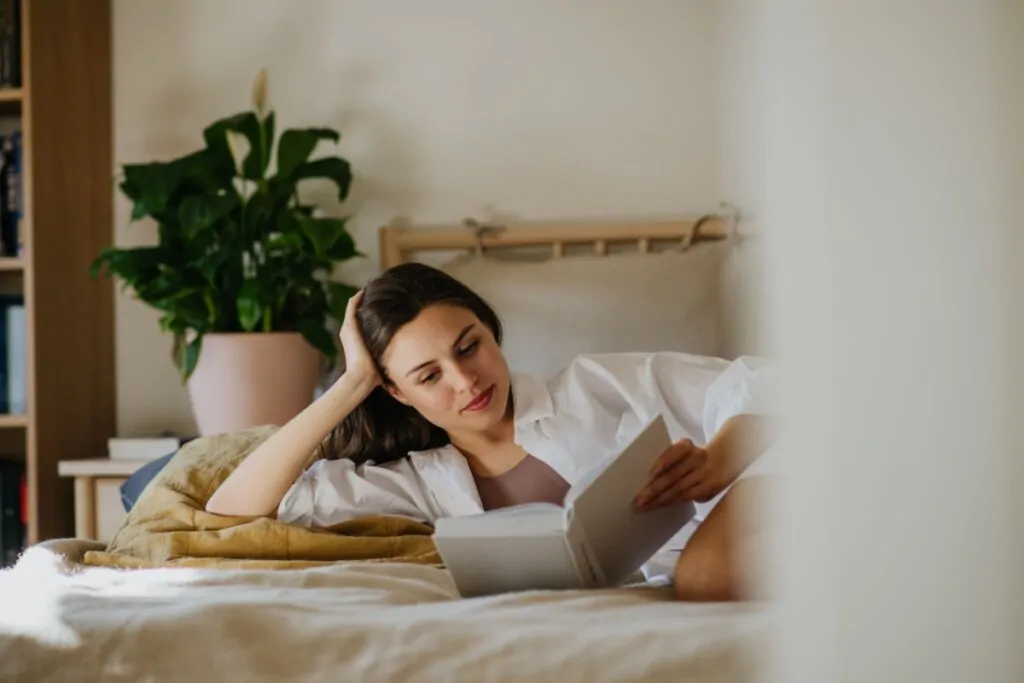 Mulher com o cabelo longo solto, usando camisa branca de botões e regata marrom deitada na cama lendo um livro