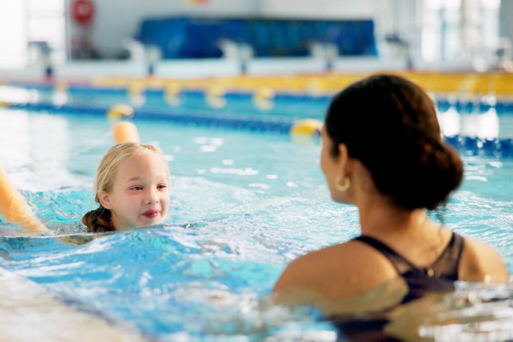 Criança loira em aula de natação com instrutora, em piscina coberta