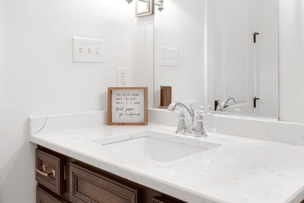 Pia de banheiro branca em móvel em madeira com placa de decoração com frase no canto da pia 