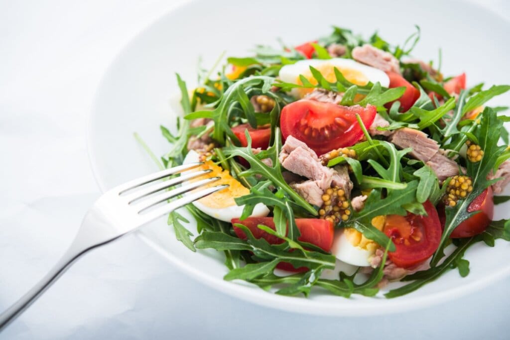 Salada de atum com ovo, rúcula e tomate servido em prato branco em cima de mesa branca com garfo ao lado