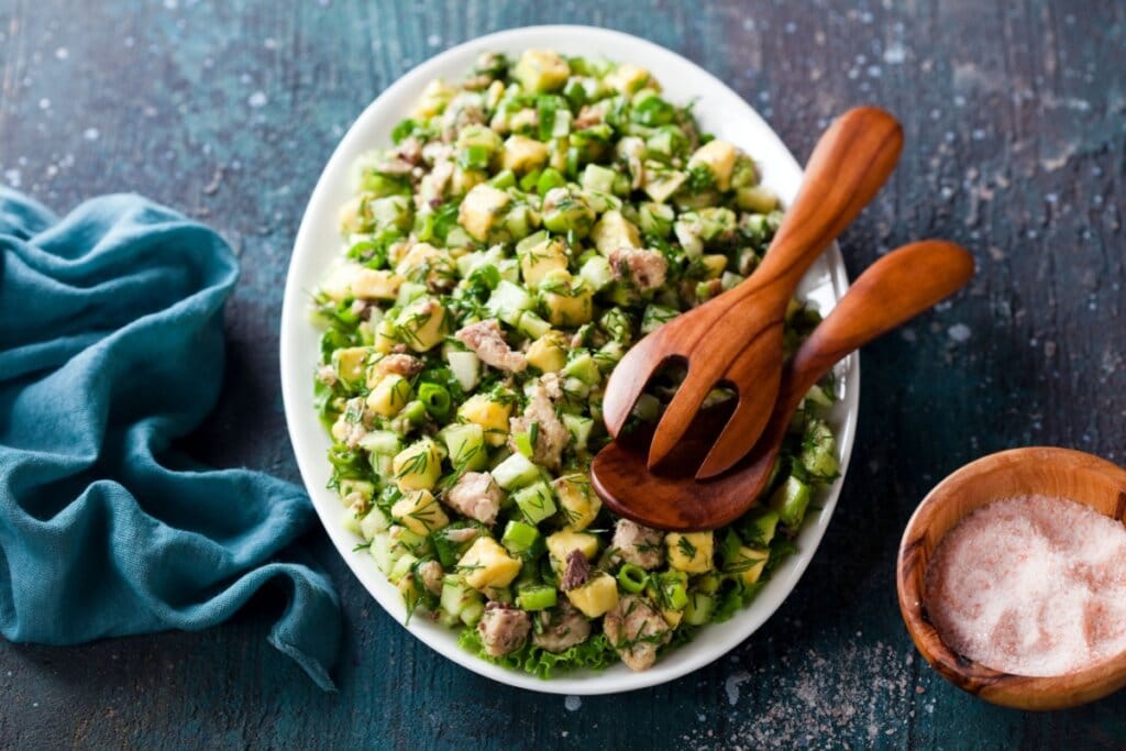 Salada de sardinha com abacate e pepino servida em tigela de cerâmica branca com colheres de pau em cima, em cima de mesa de pedra