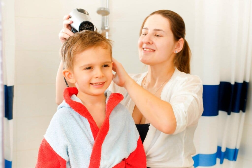 Mãe secando o cabelo do filho com secador após atividade aquática, enquanto o menino sorri usando um roupão de banho