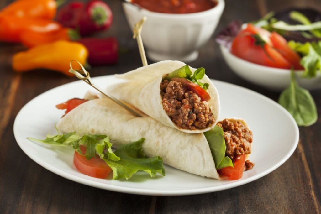 Dois burritos de carne moída em um prato branco; ao redor, estão potes com os ingredientes