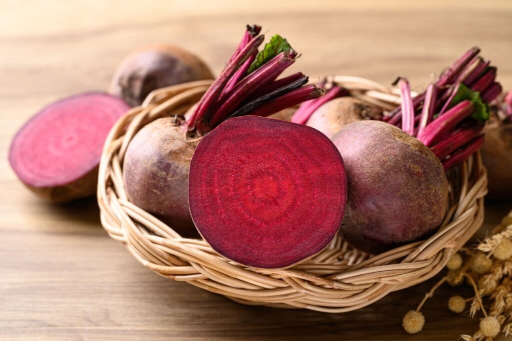 Cesto de madeira com beterrabas, uma delas partida ao meio em cima de mesa de madeira, com uma metade de beterraba e outra inteira ao lado do cesto