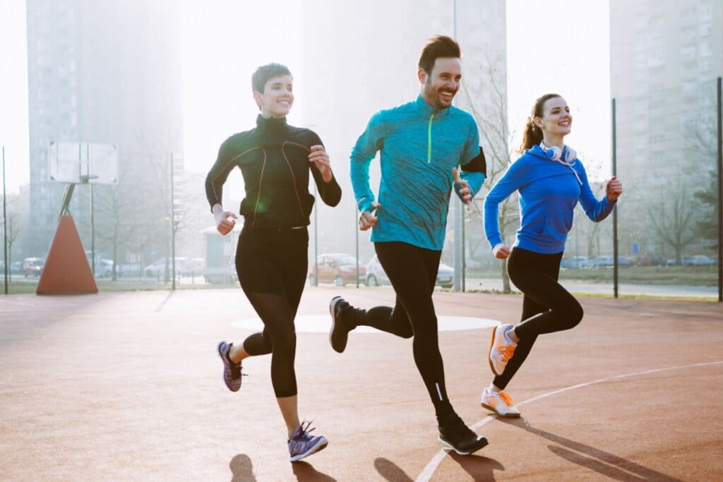 Três pessoas correndo ao ar livre, sorrindo e usando roupas esportivas de frio