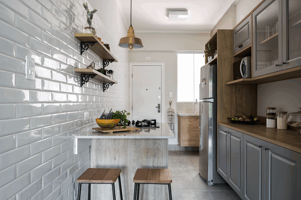 Cozinha com armários em madeira e portas na cor cinza, com bancada de mármore com cooktop e espaço para comer com dois bancos de ferro com assento em madeira e prateleiras na parede de piso branco