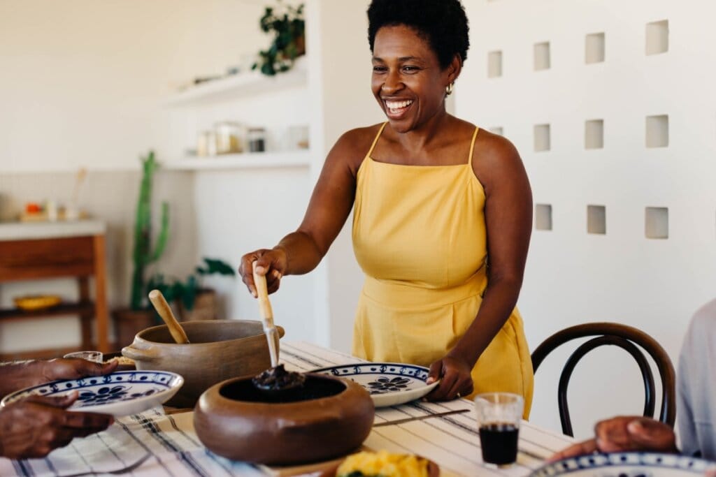 Mulher usando vestido amarelo, sorrindo e colocando feijoada em um prato 
