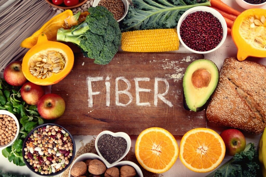Mesa de madeira, onde está escrito fiber com giz branco e diversos alimentos ricos em fibras como laranja, abacate, milho, maçã e outros