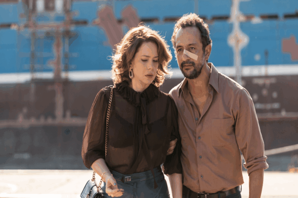 Imagem de um casal andando junto na rua, a mulher está usando uma camisa de manga longa de tule e um top preto e o homem usando uma camisa de botões marrom e com um curativo no rosto