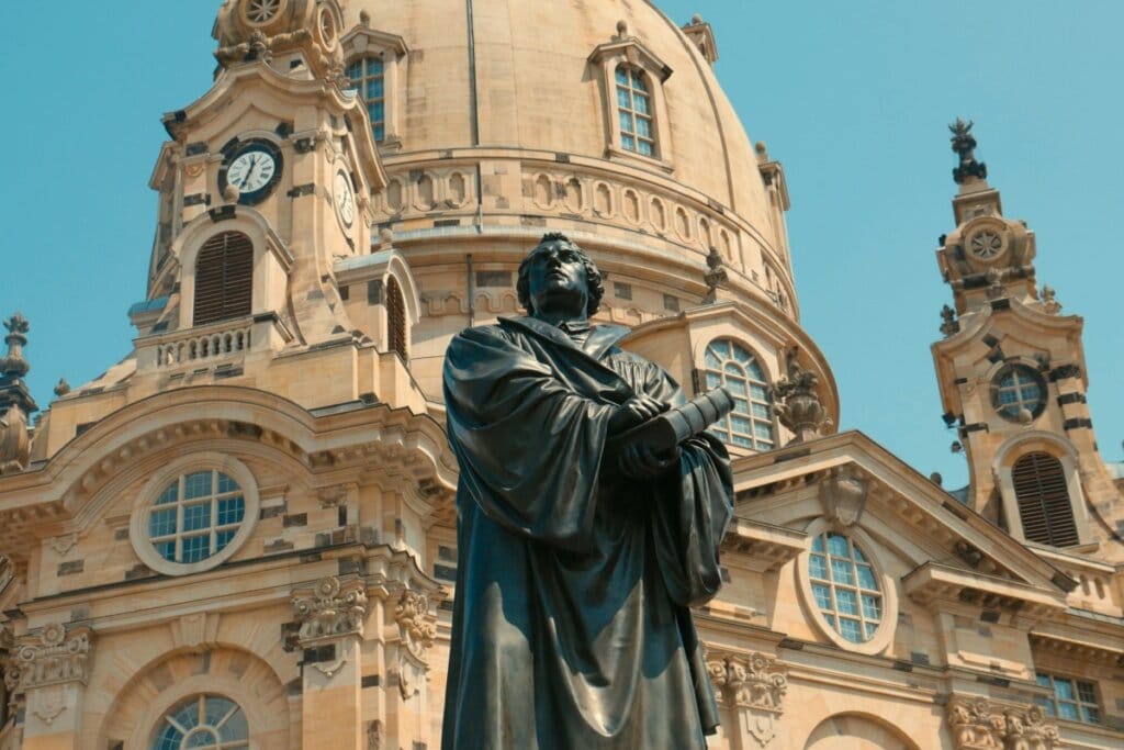 Estátua de um homem com livros nas mãos e ao fundo há uma igreja 