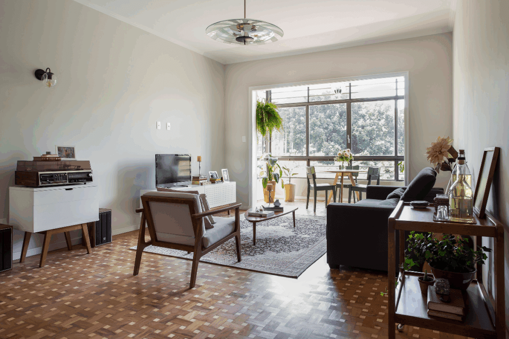 Sala com piso de taco, sofá preto e cadeiras em madeira com estofado branco e móvel com TV branco e paredes pintadas em tons neutros e com janela ampla ao fundo