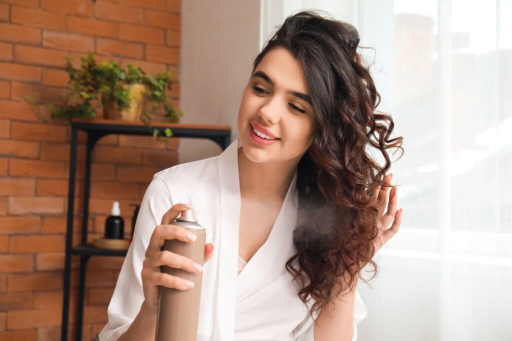 Jovem com o cabelo cacheado passando spray nos cachos do cabelo, ela está usando um roupão branco