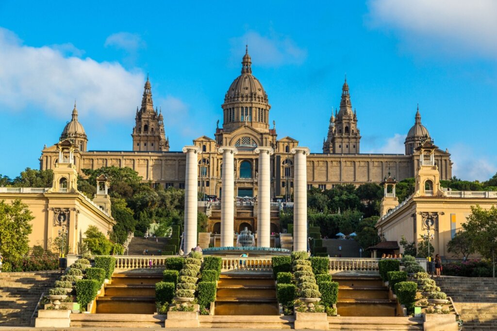 Imagem da entrada do Museu Nacional d’Art de Catalunya (MNAC) com escadas e plantas nas laterais e  4 pilastras e prédio ao fundo