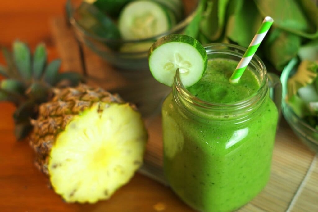 Caneca de vidro com suco de pepino com abacaxi e espinafre em cima de uma mesa com pedaços de abacaxi, pepino e folhas de espinafre 