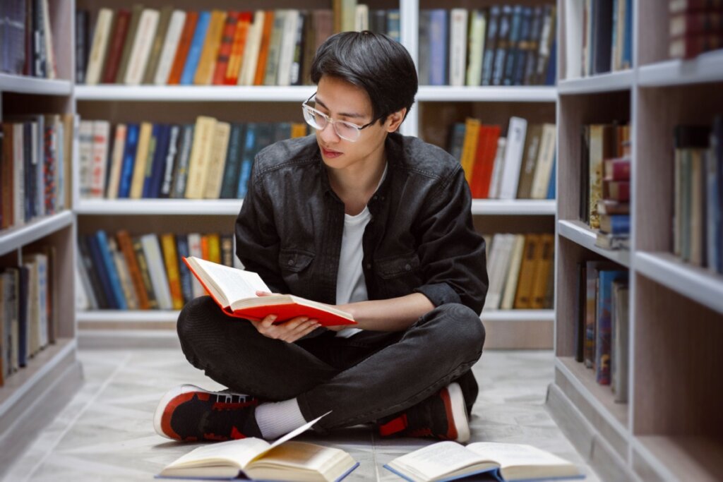 Jovem sentado no chão de uma biblioteca lendo livro