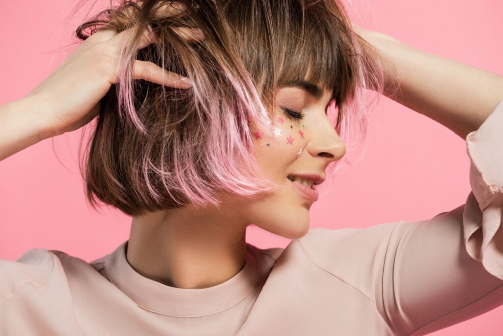mulher com cabelo curto castanho com mechas rosas, com estrelas no rosto, olhos fechados e mãos no cabelo