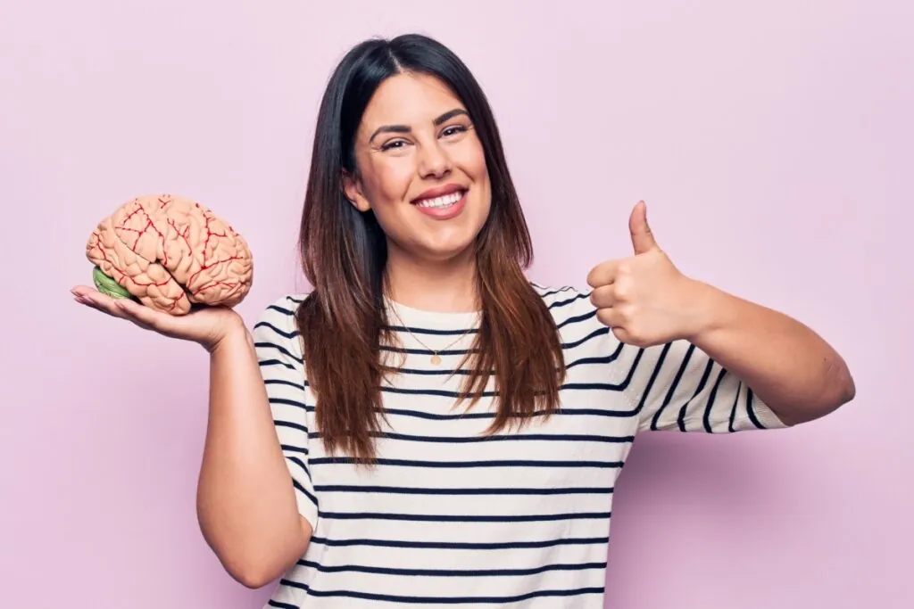 mulher de cabelo longo, sorrindo, fazendo "joia" com o dedo e segurando cérebro na mão