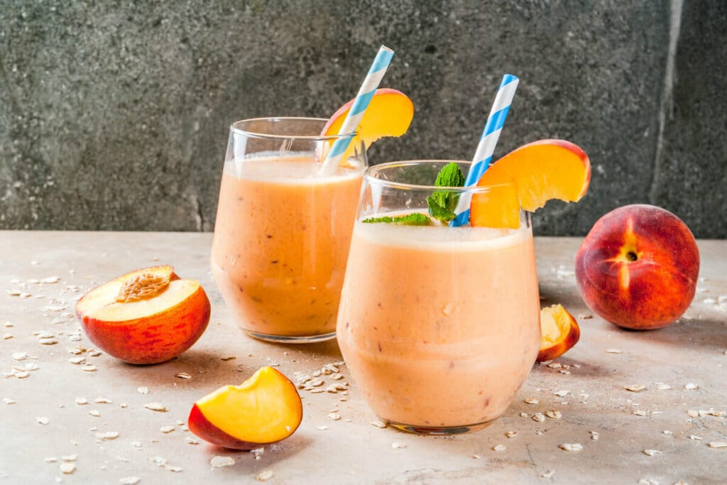 Dois copos de vidro com vitamina de pêssego em cima de uma mesa de madeira com flocos de aveia e pedaços de pêssego