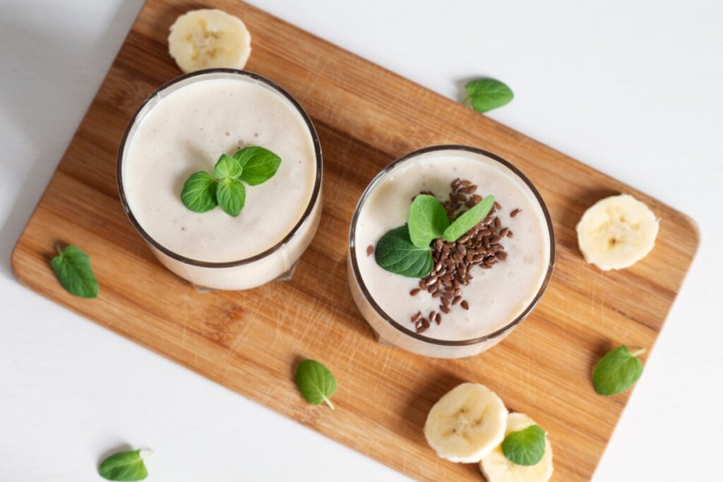 Vitamina de banana com linhaça servida em copos de vidro em cima de tábua de madeira com rodelas de banana e folhas de hortelã ao redor