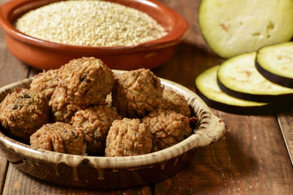Almôndegas de abobrinha com quinoa em um recipiente bege, sobre uma mesa de madeira, acompanhadas de grãos de quinoa e fatias de abobrinha