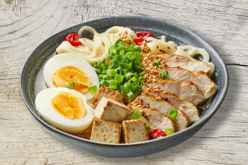 Tigela de lámen japonês com caldo claro, pedaços de frango grelhado, cubos de tofu, ovos cozidos, cebolinha e sementes de gergelim sobre superfície de madeira clara