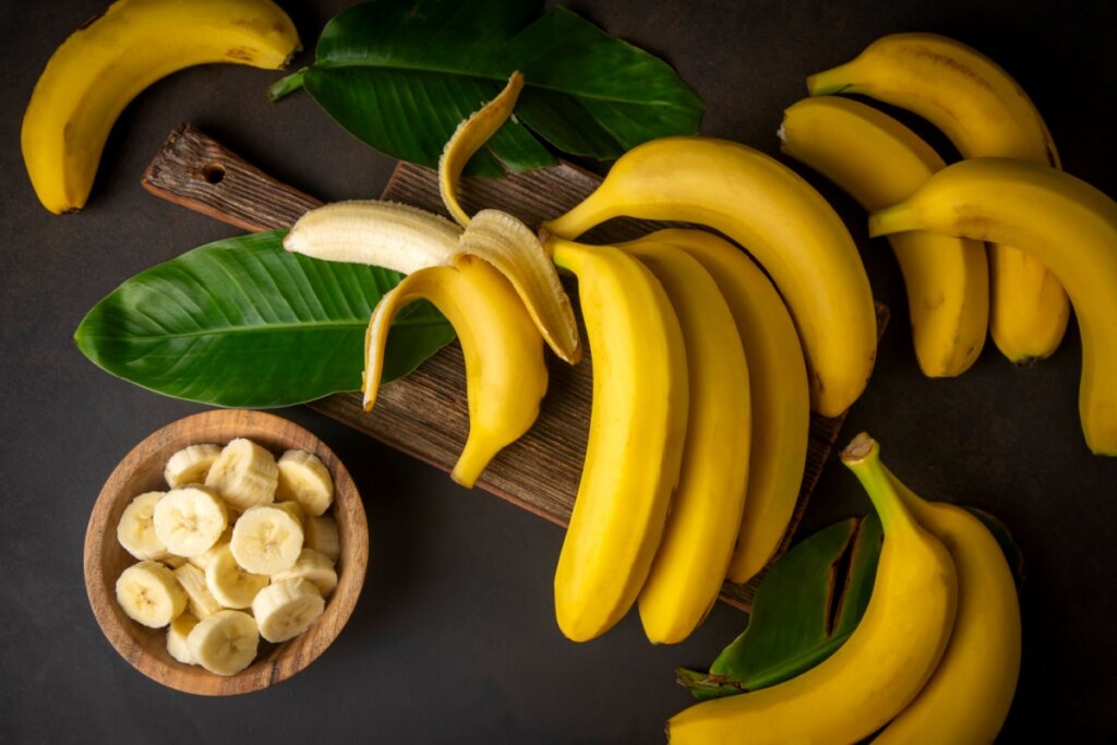 bananas com casca amarela em cima de tábua com folhas verdes e pote de madeira com rodelas de banana ao lado