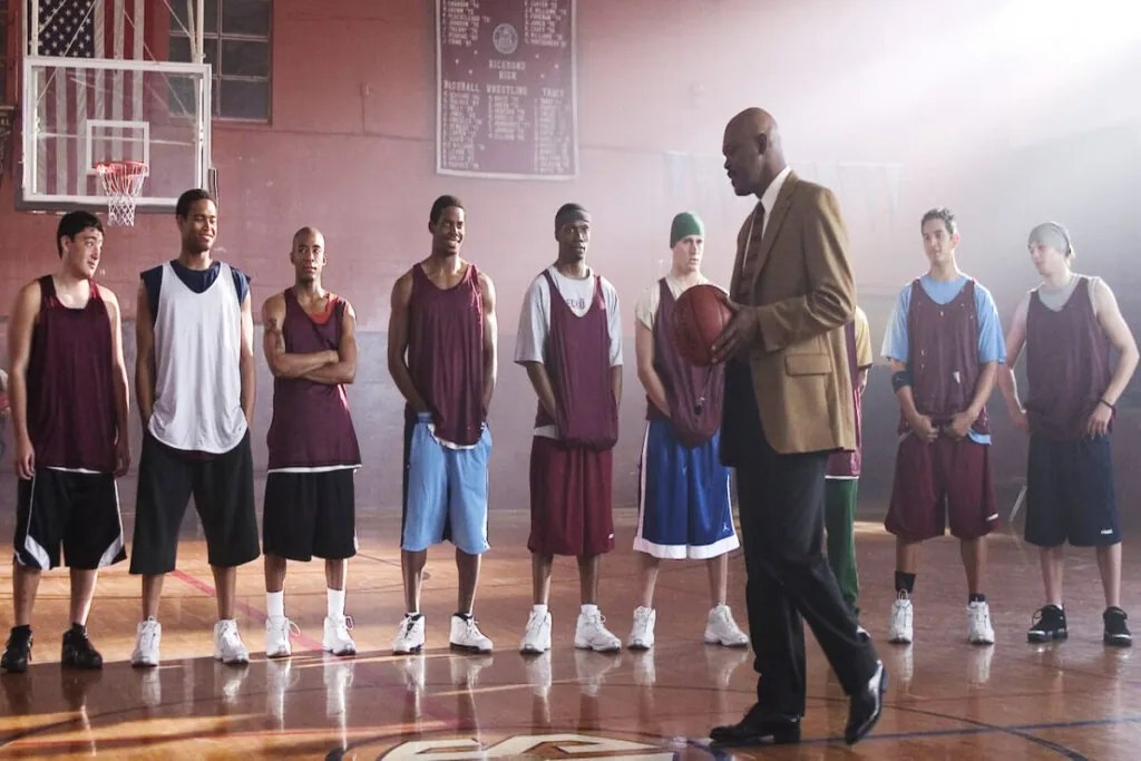 Grupo de jovens usando uniforme de time de basquete em quadra de escola recebendo orientações de treinador usando roupas sociais marrom e preta 