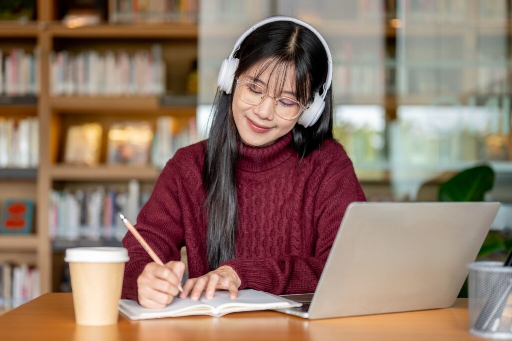 Jovem com cabelo solto, liso e de franja usando fone de ouvido cinza, blusa de lã vermelha escrevendo em caderno com notebook ao lado