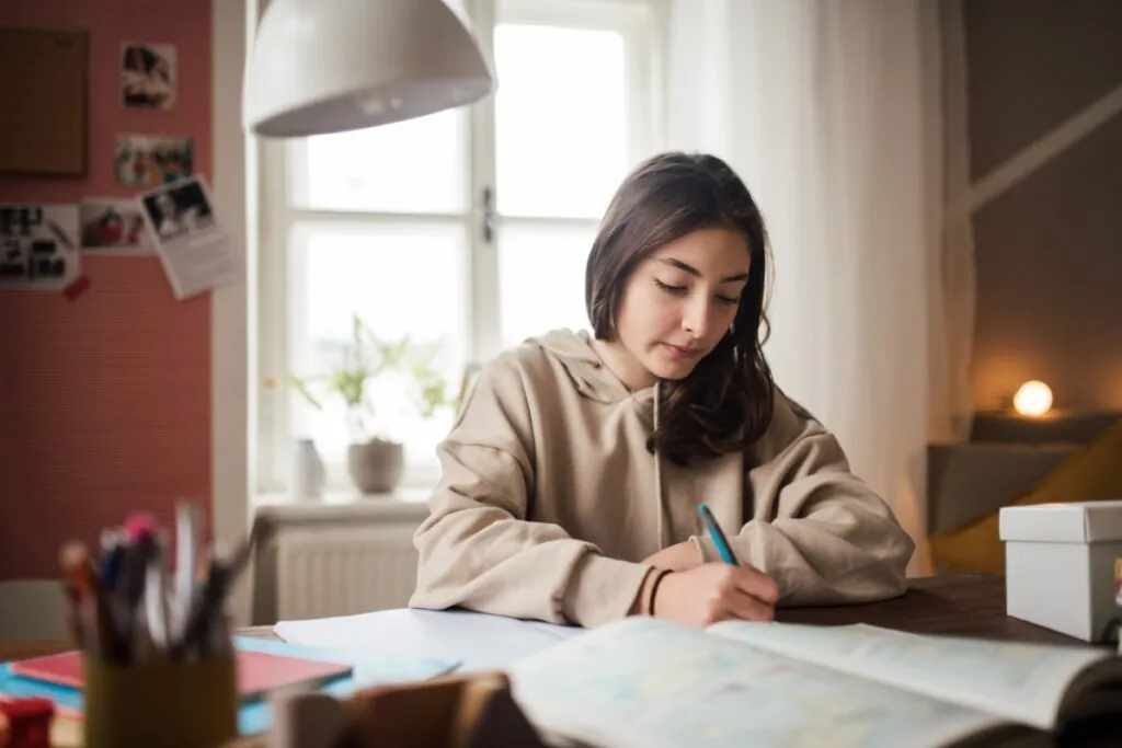 Jovem com cabelo liso, na altura do ombro, usando moletom bege, escrevendo em caderno estudando