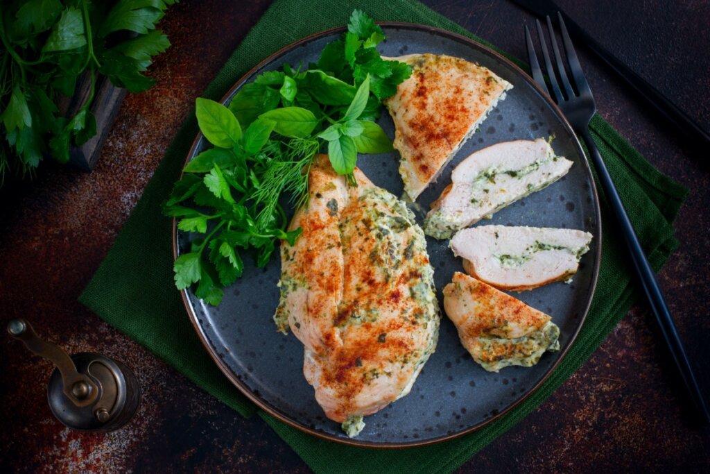 prato azul com peito de frango assado e, ao lado, outro peito de frango assado fatiado e recheado com creme de ervas. salsinha e dill ao lado no prato